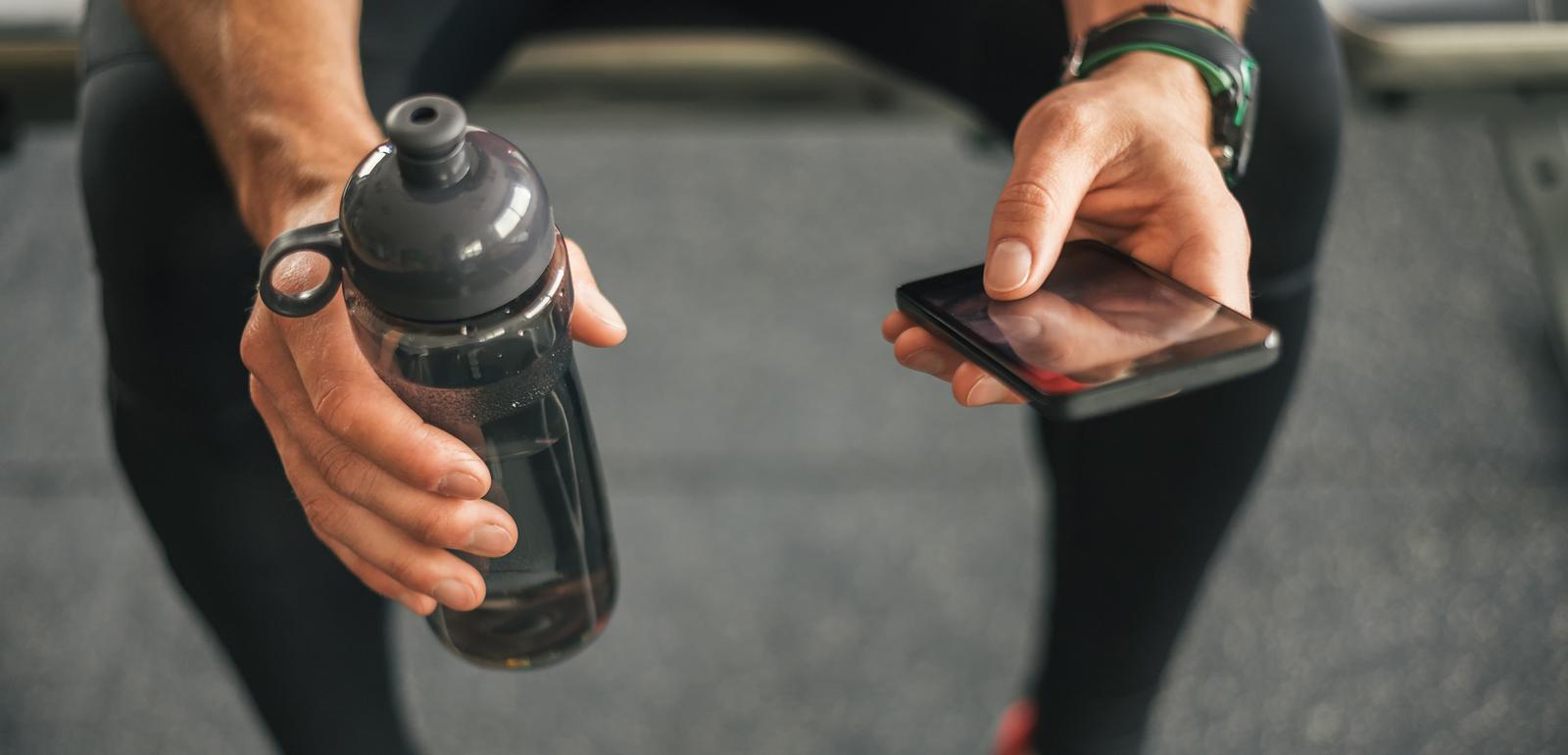 Nahaufnahme einer Hand, die eine Wasserflasche h&auml;lt, und einer anderen, die ein Smartphone in einem Fitnessstudio h&auml;lt.