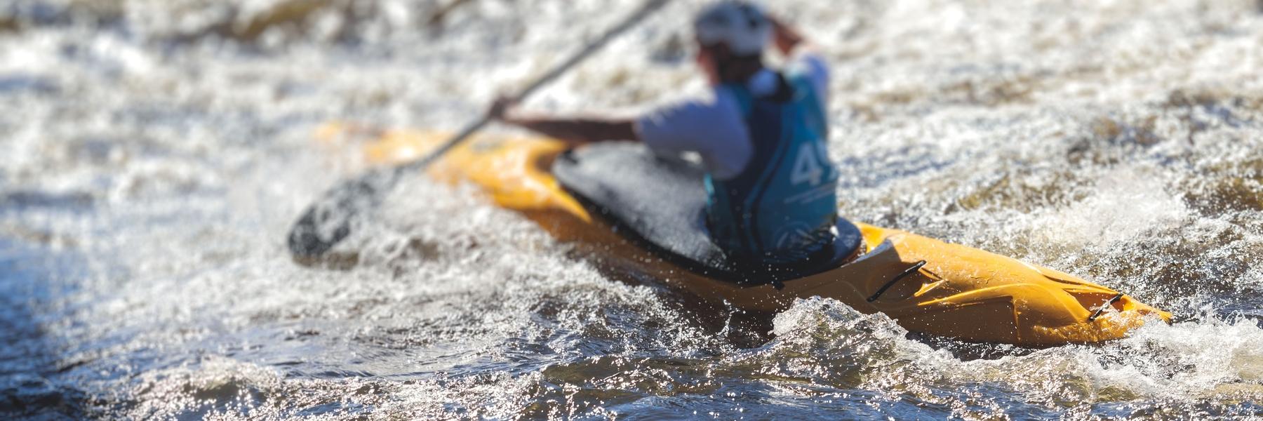 Paddler in einem gelben Kajak k&auml;mpft sich durch turbulent gewelltes Wasser.