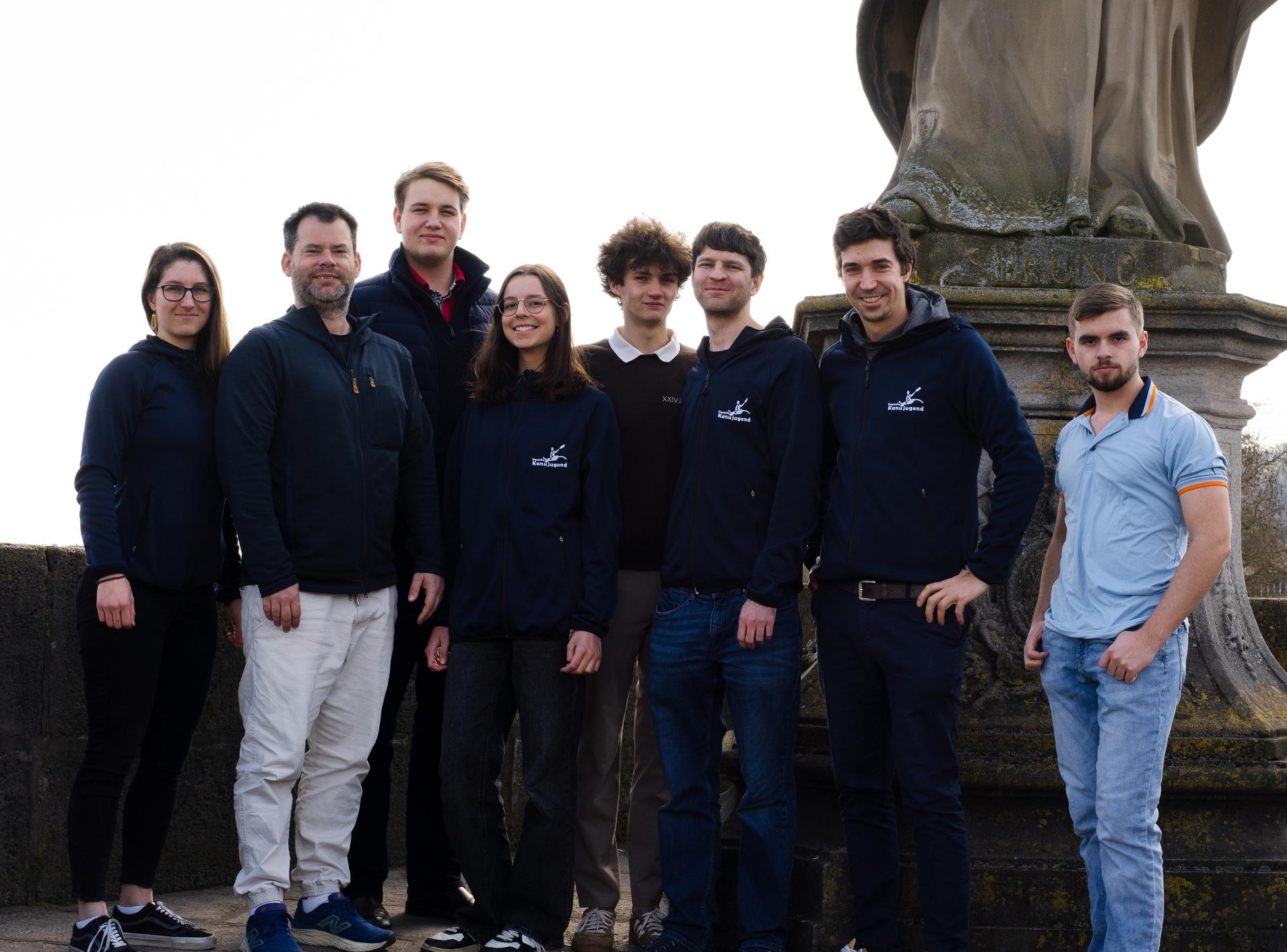 Gruppenfoto von acht Personen vor einer Statue, alle tragen verschiedene Oberbekleidung und stehen auf einer steinernen Fläche.