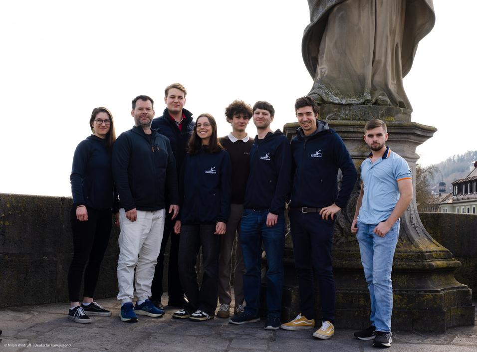 Gruppenfoto von acht Personen vor einer Statue, alle tragen verschiedene Oberbekleidung und stehen auf einer steinernen Fl&auml;che.