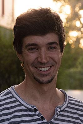 Porträt eines lächelnden Mannes mit kurzen, lockigen Haaren, getragen in einem gestreiften T-Shirt, vor unscharfem Hintergrund.