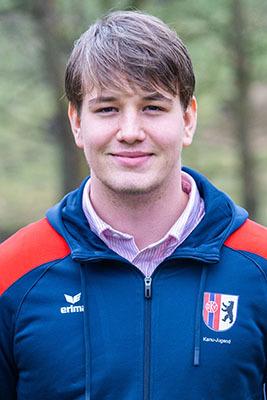 Junger Mann in blauer Sportjacke mit rotem Kragen lächelt vor verschwommenem, natürlichem Hintergrund.