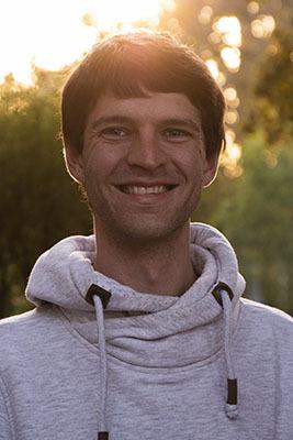 Lächelnder Mann in grauem Hoodie vor unscharfem Hintergrund mit Sonnenlicht durch Bäume.