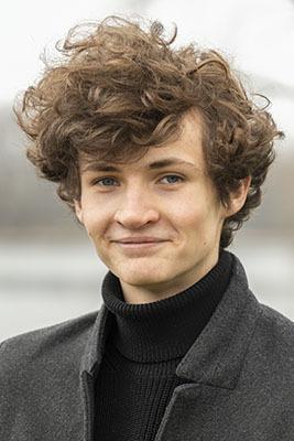 Junger Mann mit lockigem, braunem Haar, trägt einen schwarzen Pullover und einen grauen Mantel, Hintergrund unscharf.