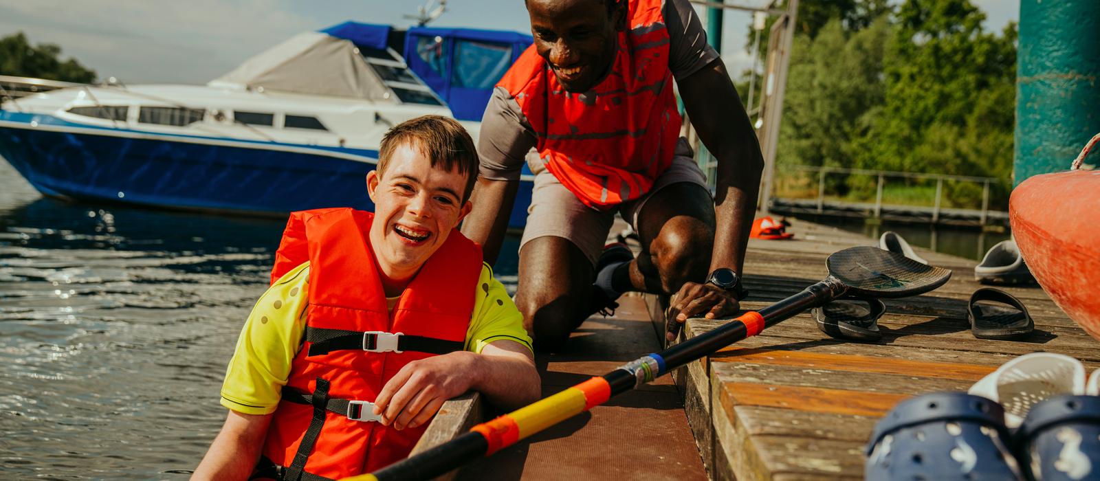 Zwei Personen in Rettungswesten lachen am Ufer eines Gewässers, während sie ein Paddel und einen Kajak im Hintergrund haben.